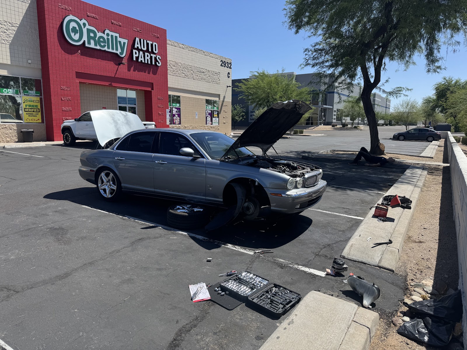 Silver Jaguar sedan with its hood up and front wheel off, mid repair in an O'Reilly Auto Parts parking lot under a clear blue sky